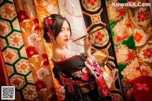 A woman in a kimono sitting on a red carpet holding a fan.